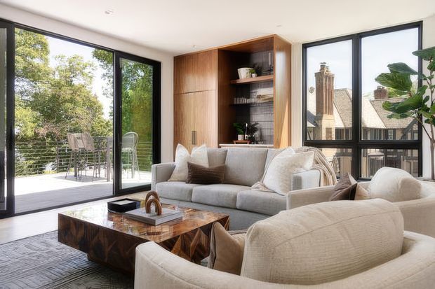 Bright contemporary living room with large black-framed glazed doors and windows, opening to an outdoor terrace. The room features neutral upholstered sofas and armchairs, a dark patterned timber coffee table, a full-height warm timber built-in shelving and cabinetry wall, and views toward neighbouring traditional buildings and greenery.