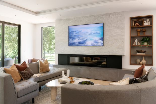 A bright, refined contemporary living room centered around a large linear fireplace set within a textured light stone or plaster feature wall. A wall-mounted television sits above the fireplace, with built-in dark timber shelving displaying books, ceramics and greenery to the side. Curved and modular neutral upholstered seating surrounds a pale round coffee table, while expansive windows bring in views of greenery.