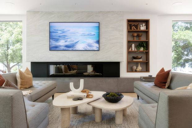 Contemporary living room with a symmetrical built-in fireplace wall, pale textured stone or plaster surround, wall-mounted television, timber display shelving, and large windows overlooking greenery. Two light upholstered sofas face a pair of rounded pale coffee tables, creating a bright, refined seating area.