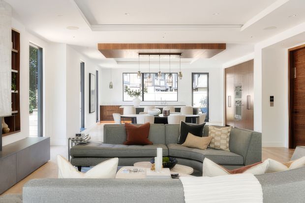 Bright, refined open-plan living and dining area with a large grey sectional sofa in the foreground, white and neutral upholstery, warm timber built-in cabinetry and ceiling detailing, and expansive black-framed windows. The room has a clean contemporary fit-out with recessed lighting, a sculptural linear pendant above the dining table, and a calm neutral palette with rust and black accent cushions.