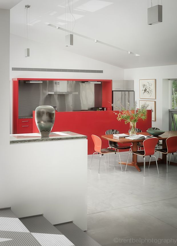 A bright, open-plan modern kitchen and dining area with a dramatic red kitchen surround and island cabinetry, stainless-steel splashback and appliances, white walls and ceiling, and a pale tiled floor. A round wooden dining table is surrounded by red and dark chairs, with pendant lights above and a sculptural vase on the island. Stairs descend in the foreground, and a window provides greenery at the right.