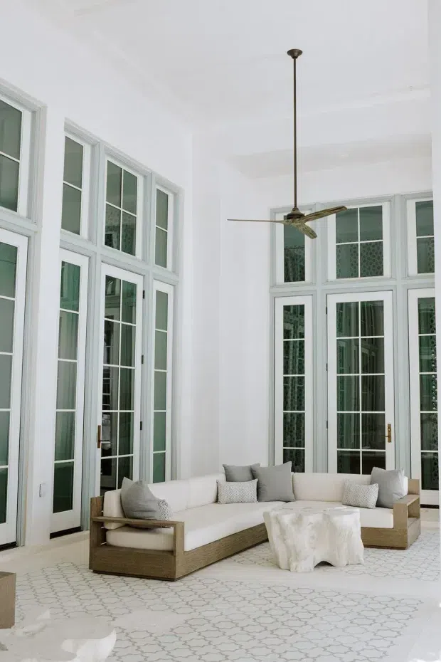 Bright, airy sunroom or living space with very tall white-framed multi-pane French doors and windows, a high white ceiling, and a contemporary ceiling fan. A large light-toned sectional sofa with woven wood-look bases and neutral cushions sits on a pale patterned floor, creating a relaxed indoor-outdoor feel.
