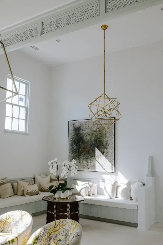 Bright, refined sitting room with a built-in window seat running along the corner, layered neutral cushions, patterned upholstered lounge chairs, and a dark round occasional table topped with orchids. White walls and ceiling are enhanced by decorative perforated ventilation grilles, tall multi-pane windows, a geometric brass pendant light, and a large abstract artwork. Strong natural light creates an airy yet elegant atmosphere.