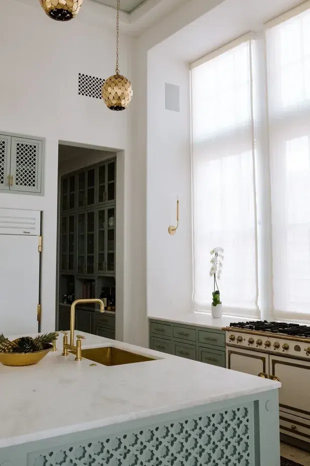 Bright, tall-ceilinged kitchen with a large white island featuring a decorative perforated geometric panel, integrated brass sink fixtures, pale stone benchtop, muted green-grey cabinetry, and a classic freestanding range. Full-height sheer window coverings provide abundant natural light. Dark built-in pantry cabinetry, patterned ventilation grilles, and gold pendant lights add decorative character.