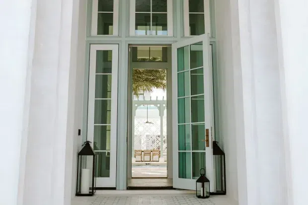 A tall, elegant white architectural entrance with pale sage-green framed French doors and transom windows. One door is open, revealing a bright covered outdoor courtyard or terrace beyond with patterned screens, palm foliage, and seating. Black lanterns sit on the tiled threshold, creating a refined, airy entry composition.