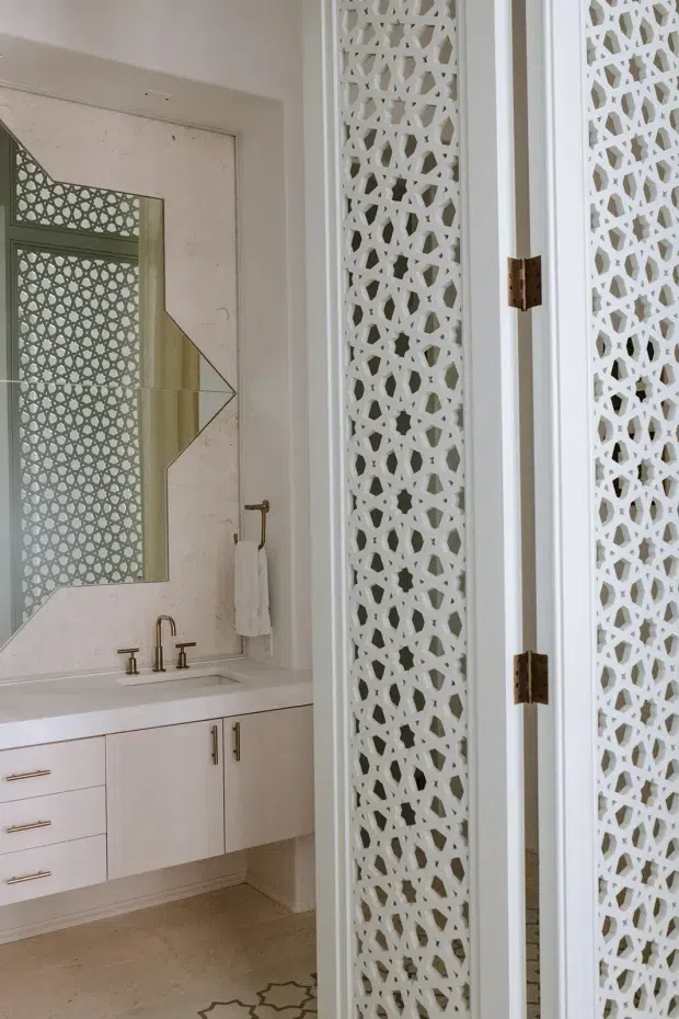 A refined bathroom vanity area viewed through partially open white decorative doors or screens. The room features a wall-mounted white vanity with drawers and cupboards, a white benchtop, brushed brass or bronze tapware and hardware, a large geometric-framed mirror, and intricate white patterned breeze-block-style screens that are also reflected in the mirror. The palette is predominantly white and off-white with warm metallic accents and a restrained geometric aesthetic.