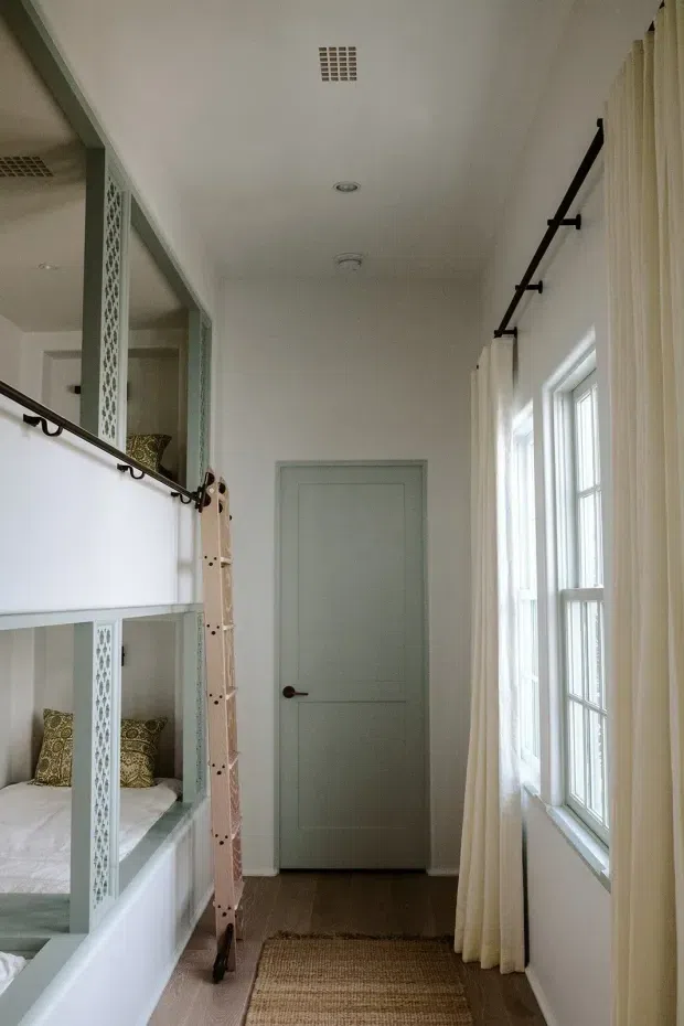 A narrow, light-filled bedroom corridor with built-in bunk beds on the left, pale decorative perforated panels, patterned pillows, a wooden ladder, and a pale green interior door at the end. Tall white-framed windows with full-length cream curtains line the right wall. Dark timber flooring and a natural woven runner add warmth to the otherwise soft white and sage palette.