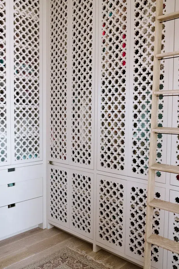 A bright interior storage and wardrobe area featuring extensive white built-in cabinetry with intricate geometric lattice or fretwork doors. A pale timber ladder leans against the cabinetry, while light timber flooring and the edge of a patterned rug add warmth. The decorative perforated panels create a refined, highly detailed feature wall.