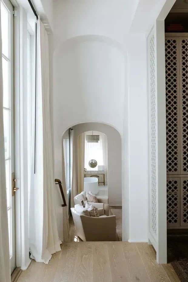 A bright, refined interior hallway or landing with tall white-framed windows and sheer curtains on the left, pale timber flooring, and a sequence of arched openings leading toward a softly furnished sitting area. A decorative patterned screen or tiled wall is visible on the right, while the distant room contains neutral upholstered chairs, a small table, and a pendant light. The space has classical architectural detailing, high ceilings, and a calm, elegant neutral palette.