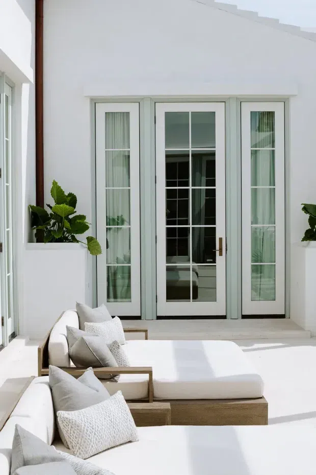 Bright, airy white courtyard-style living space with two low-profile white upholstered sofas or daybeds arranged in front of tall French doors. The doors have pale painted frames, divided glass panes, and sheer curtains, opening visually toward a darker interior. White rendered walls, pale flooring, light timber sofa bases, neutral cushions, and potted greenery create a calm contemporary coastal feel.