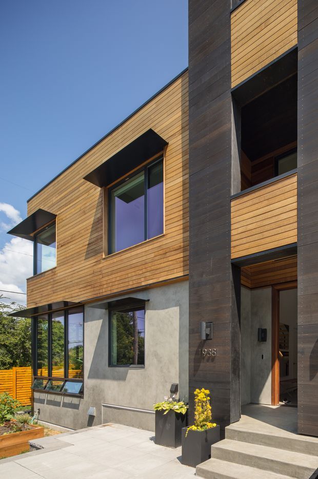 Exterior view of a contemporary multi-level residence with warm horizontal timber cladding, dark vertical timber-clad structural elements, and grey rendered lower walls. Large glazed windows and projecting black sunshades define the facade. A recessed entry is reached by broad concrete steps, with paved landscaping and planters in the foreground.