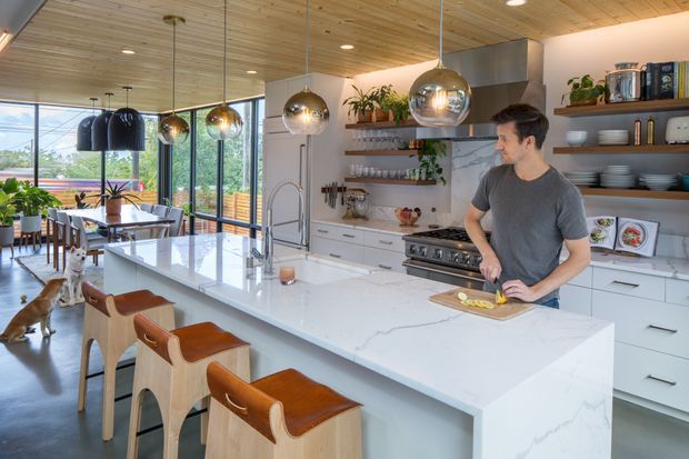 Bright, spacious open-plan kitchen with a large white stone island, integrated seating, white cabinetry, open timber shelving, and a timber-lined ceiling. Multiple pendant lights hang above the island, while full-height glazing connects the kitchen to a leafy outdoor setting and dining area. A person is preparing food at the island, and a dog is present in the background.