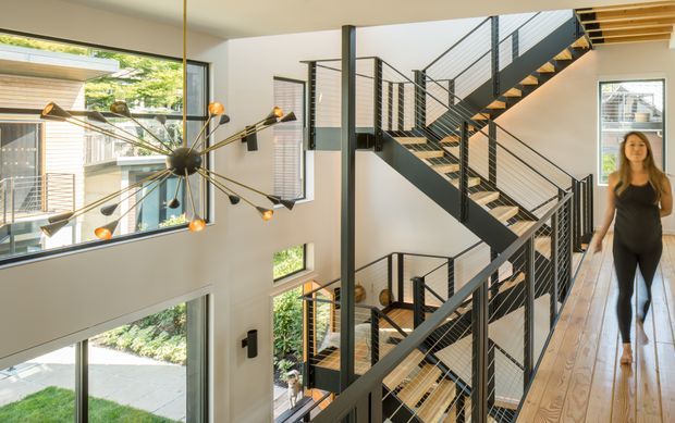 A bright multi-level interior stair hall with an open industrial-style black metal staircase and railings, light timber floors, white walls, extensive glazing, and a sculptural multi-arm pendant light. The space appears to connect several levels around a tall open void, with exterior greenery visible through the windows.