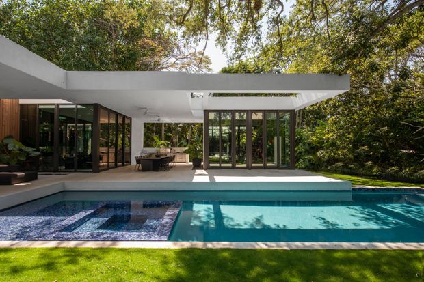 A modern single-storey residence viewed from the pool, with a crisp white flat-roofed pavilion surrounding a covered outdoor terrace. Extensive dark-framed floor-to-ceiling glazing opens the living spaces to the pool and lush garden, while a rectangular roof opening brings light into the terrace. The foreground features a geometric tiled pool with a shallow sun shelf and strong reflections of the surrounding trees.