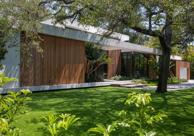A low-slung contemporary residential exterior set within a landscaped garden. The facade combines warm vertical timber cladding with white and grey horizontal roof and wall planes, expansive dark-framed glazing, and a recessed entry walkway. Mature trees and lush planting surround the building, with a manicured lawn in the foreground.