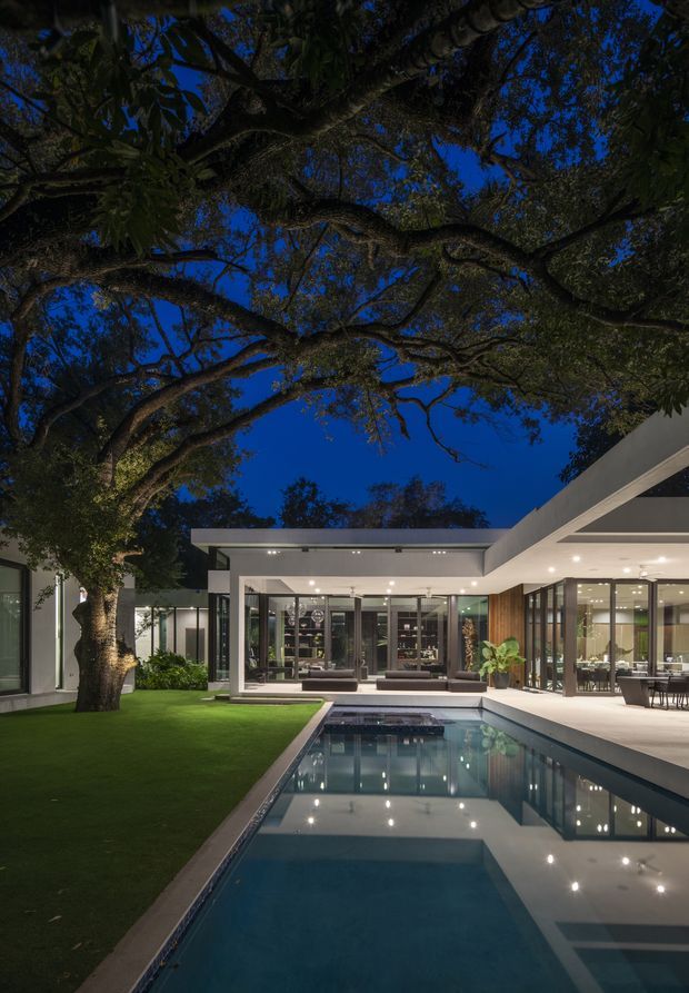 Twilight exterior view of a refined modern residence arranged around a rectangular swimming pool and manicured courtyard lawn. The house features crisp white rendered walls, extensive floor-to-ceiling glazing in dark frames, strong horizontal flat-roof forms, recessed downlighting, and an open covered outdoor dining area. A large mature tree forms a dramatic canopy over the courtyard, while the pool reflects the illuminated architecture and deep blue evening sky.