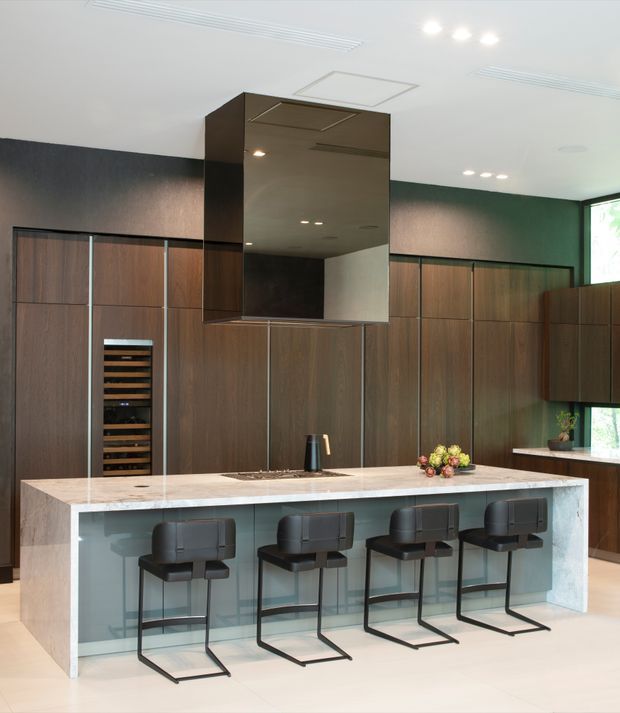 Contemporary kitchen with a large central island, pale stone benchtop and waterfall ends, four dark bar stools, extensive dark timber cabinetry, integrated wine storage, and a prominent mirrored rangehood enclosure. The room has a clean, refined fit-out with recessed lighting and a dark green and timber-toned palette.