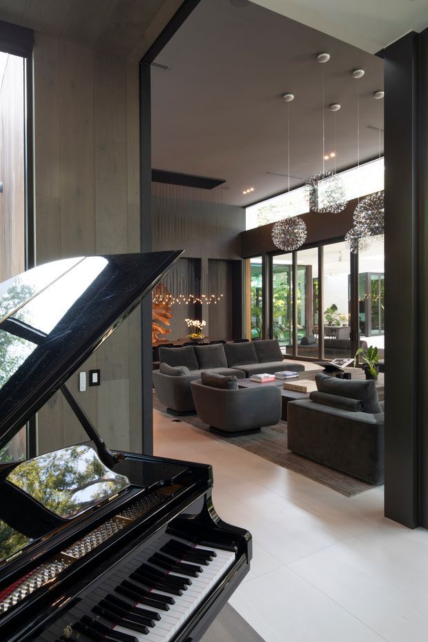 A sophisticated contemporary living room viewed from beside a glossy black grand piano. The space features dark charcoal sofas and armchairs arranged around a low coffee table, pale large-format flooring, vertically grooved dark wall panelling, extensive floor-to-ceiling glazing, and multiple spherical pendant lights. An adjoining landscaped outdoor area is visible through the glass doors.