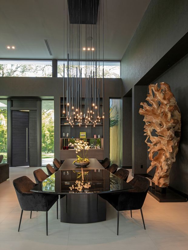 A dramatic contemporary dining room with a large glossy black dining table surrounded by upholstered dining chairs. A striking cluster of suspended pendant lights hangs over the table, extending from the high ceiling. Dark textured walls, built-in shelving, expansive clerestory glazing, a tall entry door, and a sculptural timber installation create a luxurious, moody interior.