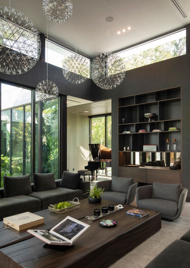 A sophisticated double-height living room with dark charcoal walls, extensive floor-to-ceiling glazing, and a large built-in display and media cabinet. A dark sectional sofa and two lounge chairs surround a substantial timber coffee table. Sculptural spherical pendant lights hang at varying heights, while a grand piano is visible in the adjoining glazed area. The room has abundant natural light, strong vertical proportions, and a refined contemporary feel.
