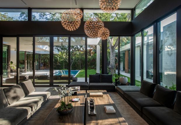 A spacious, contemporary living room with floor-to-ceiling black-framed glazing on multiple sides, opening views to a landscaped courtyard and swimming pool. The room has a high ceiling with clerestory windows, dark sofas, a large timber coffee table, and several spherical woven pendant lights. The dark interior frame contrasts with abundant natural light and greenery outside.