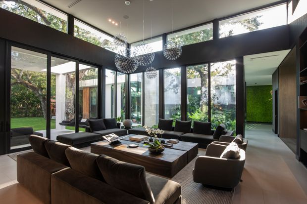 A sophisticated contemporary living room with a large dark timber sectional sofa, upholstered armchairs, and a substantial rectangular coffee table arranged around a central seating area. Floor-to-ceiling glazing and clerestory windows open the room to a leafy garden, while sculptural pendant lights hang from a high ceiling. Dark built-in shelving and wall bands contrast with pale flooring, and a vivid green living wall is visible through the adjoining opening.