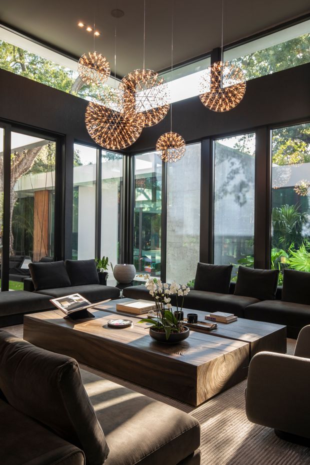 A sophisticated living room with floor-to-ceiling black-framed glazing opening toward a landscaped garden. The room features several sculptural spherical pendant lights, dark sofas and armchairs, a large timber coffee table, a textured rug, and minimalist decorative accessories. Strong natural light enters through the extensive glazing, contrasting with the dark interior finishes.