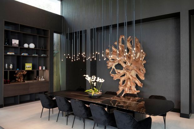 A dramatic, sophisticated dining room with a long dark dining table surrounded by upholstered dining chairs. A sculptural pendant installation with numerous hanging bulbs spans the table, while a large organic wood sculpture forms a feature display against a dark textured wall. Built-in dark timber shelving occupies the left wall, and pale flooring provides contrast. Tall glazing and a high ceiling contribute to the spacious, gallery-like atmosphere.