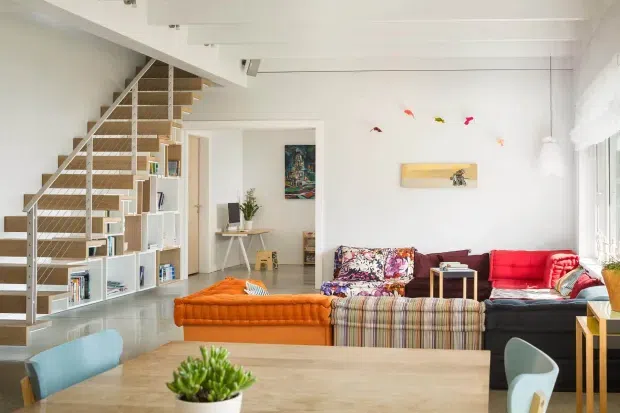 Bright open-plan living and dining area with a sculptural open-riser staircase, integrated white shelving, exposed white ceiling beams, and large windows. The room contains a colourful layered seating arrangement with an orange ottoman, patterned and striped upholstery, a red sofa, pale timber dining table, and eclectic artwork and decor. The staircase and high white interior create a contemporary, airy setting.