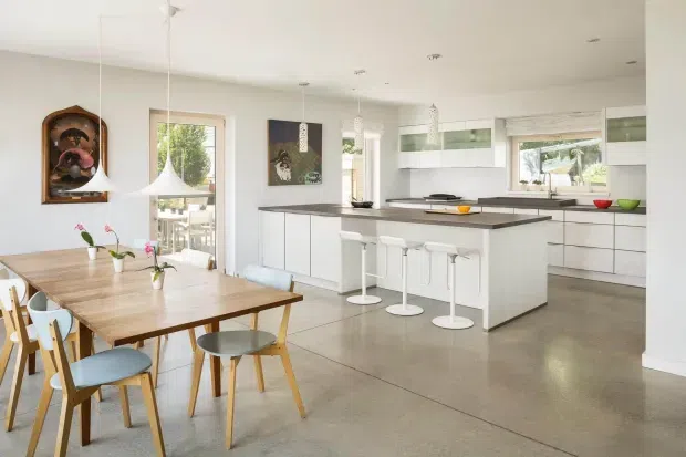 Bright open-plan kitchen and dining area with white flat-panel cabinetry, a long dark benchtop island, integrated appliances, pendant lighting, and a timber dining table with light upholstered chairs. Large windows and glazed doors provide abundant natural light, while the polished concrete floor creates a clean contemporary setting.