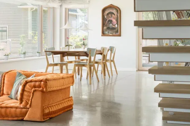 Bright open-plan interior featuring a dining area and adjacent living space. A timber dining table with mixed wooden chairs sits beside large windows, while an orange upholstered sofa occupies the foreground. Polished light concrete flooring, white walls and ceilings, and a floating timber stair with open risers define the space. The room includes a framed decorative artwork and abundant natural light, creating a warm mid-century-inspired atmosphere.