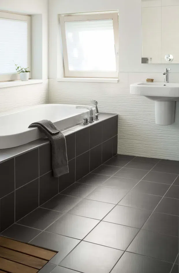 Bright, clean contemporary bathroom featuring a white bathtub with chrome fixtures, a wall-mounted white basin, dark charcoal tiled bath surround and floor, textured white wall tiles, pale cabinetry, and frosted windows with white frames. A small timber floor section is visible at the foreground threshold.