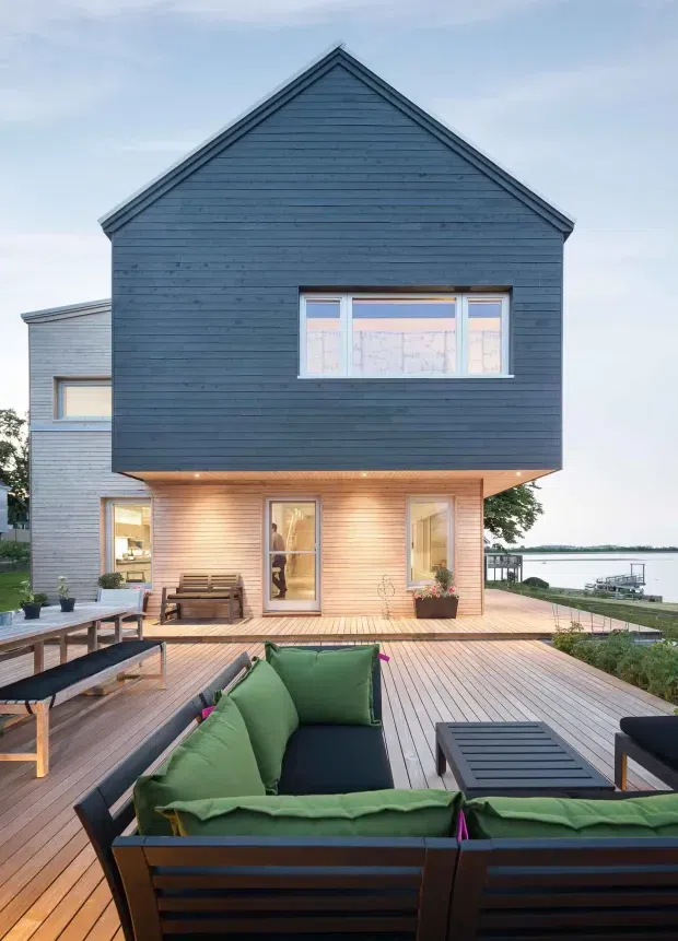 Exterior rear view of a two-storey waterfront home with a steep gabled upper volume clad in dark horizontal timber weatherboards and a lighter timber-clad ground floor. The house opens onto a broad timber deck furnished with outdoor lounge seating and a dining table, with water visible beyond the property.
