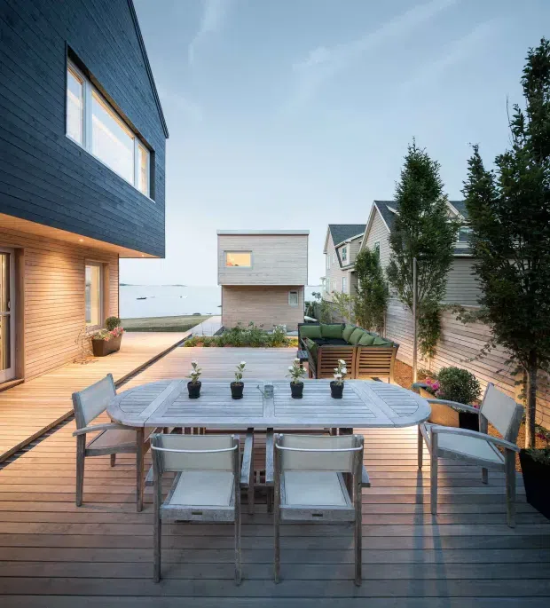 A contemporary waterfront residential terrace viewed from an outdoor dining deck. The scene features a large timber dining table with metal-framed chairs, a built-in outdoor lounge, timber decking, horizontal timber-clad houses, and potted plants. Water is visible beyond the adjoining buildings, creating an open coastal setting.