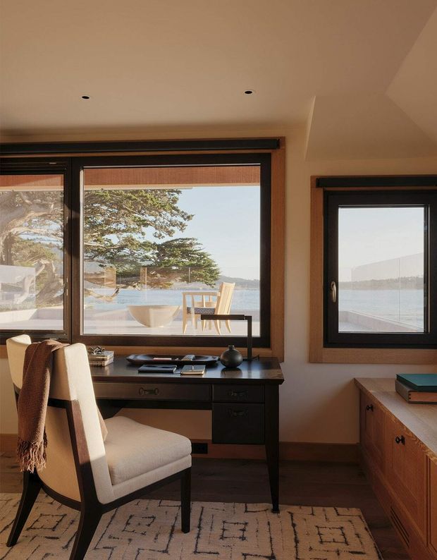 A warm, refined home office positioned beside large timber-framed windows overlooking a coastal waterfront setting. The room features a dark built-in writing desk with drawers, a cream upholstered desk chair, warm timber window surrounds and cabinetry, recessed ceiling lights, and a patterned neutral rug. Sunlight and water views create an inviting, calm atmosphere.