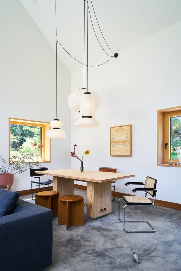 Bright, airy open-plan dining and living area with a substantial light timber dining table, cylindrical timber stools, dark blue sofa, and a black-framed cantilever chair. Multiple sculptural pendant lights hang from long exposed cords against a high white ceiling. Timber-framed windows bring in abundant greenery views, while a framed print and small vase provide restrained decoration. The space has polished concrete flooring and a clean, contemporary minimalist feel.