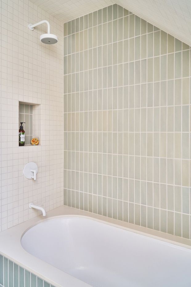 A bright, compact bathroom focused on a built-in white bathtub and shower area. The walls are tiled with small square off-white tiles, while a large feature wall uses vertically oriented pale sage-green rectangular tiles in a regular grid. White shower and bath fixtures, a recessed wall niche with toiletries, and clean white surfaces create a fresh, understated look.