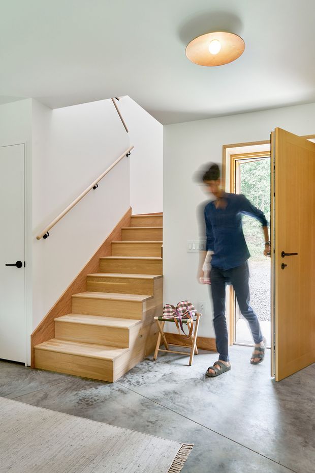 Bright residential entry hall with a timber staircase, pale wood handrail, white walls, polished concrete floor, and a timber front door opening to the exterior. A simple ceiling-mounted pendant light, narrow side window, and small folding chair complete the space; a person is captured in motion near the doorway.