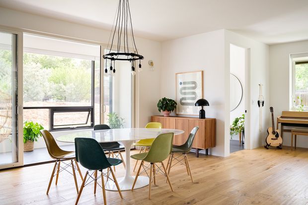 Bright open-plan dining and living area with a mid-century-inspired dining set, timber sideboard, light timber flooring, white walls, and expansive glazing opening to greenery. A sculptural pendant light hangs above the dining table, while a piano and guitar are visible in the adjoining area.