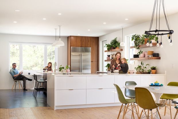 Bright open-plan kitchen and dining area with a large white island, integrated stainless-steel refrigerator, warm timber cabinetry, open display shelving, pendant and track lighting, and a dining table with colourful modern chairs. Large windows provide abundant natural light, while plants and everyday activity create an inviting family-oriented atmosphere.