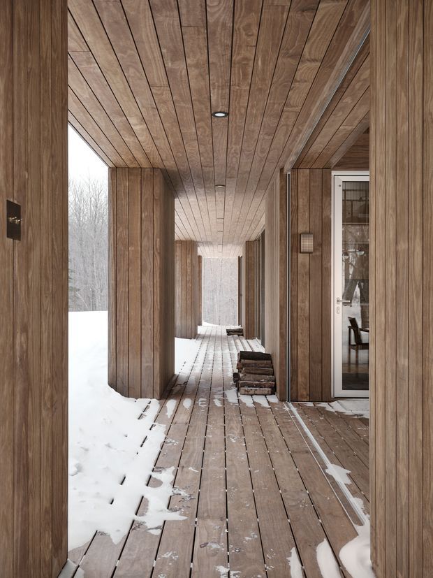 A long, covered exterior walkway or deck beside a timber-clad building in a snowy woodland setting. Vertical timber wall cladding, matching timber soffit and decking create a continuous warm wood envelope, with recessed ceiling lights, structural columns, a glazed entry door, and stacked firewood along the passage. Snow is accumulated on the deck and ground outside.