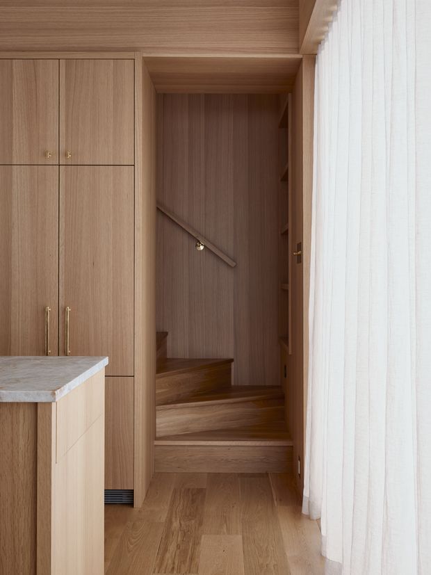 Warm, light-wood interior showing a compact stairway integrated into full-height timber cabinetry. The staircase has timber treads, a timber-lined wall and simple diagonal handrail, with open shelving tucked along the side. A kitchen island with a pale stone benchtop is visible at left, while sheer white curtains cover glazing on the right. The composition is minimalist and highly cohesive, with extensive built-in joinery and timber flooring.