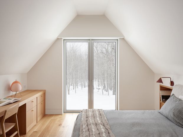 A bright attic-style bedroom with a sloping ceiling and large central glazed double doors overlooking a snowy wooded landscape. The room features pale warm timber flooring, soft off-white walls, a low timber sideboard and desk, a small wall-mounted shelf, and a neatly made bed in muted grey textiles. The design is simple, calm, and contemporary with natural materials and restrained furnishings.