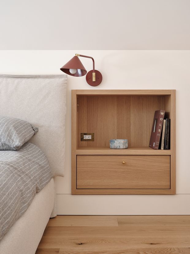 A close-up of a calm, minimalist bedroom corner showing a softly upholstered bed with grey striped bedding, a wall-mounted timber bedside cabinet with an open shelf and drawer, and a sculptural wall lamp in muted burgundy with brass detailing. The room has warm oak flooring, pale neutral walls, and refined built-in joinery.