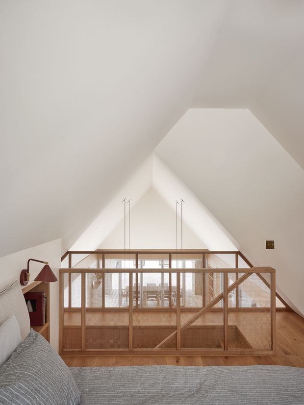 A warm, minimalist bedroom overlooking a timber-framed mezzanine or stair void. The room has a steeply pitched white ceiling with a geometric dormer-like opening, white walls, warm timber flooring, and a woven timber-and-mesh balustrade enclosing the lower level. A partially visible bed with grey textured bedding occupies the foreground, with a small wall-mounted reading lamp and built-in shelving at left. Bright daylight enters from the glazed area beyond the balustrade.