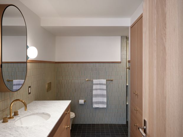 A refined bathroom with a warm, contemporary fit-out. The room features pale vertically arranged wall tiles with a timber trim, dark square floor tiles, a white marble-look vanity benchtop with integrated basin, timber cabinetry, brass tapware and towel rail, an oval wall mirror, and a timber door and tall cabinetry. A toilet and shower area are visible toward the rear.