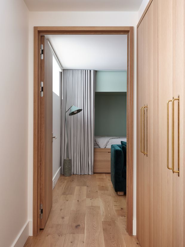 A view through a timber-framed doorway into a compact, thoughtfully designed bedroom. The room features warm timber flooring, a built-in timber wardrobe with long brass handles in the foreground, a muted green built-in sleeping nook, grey floor-to-ceiling curtains, and a teal upholstered chair. A sculptural floor lamp sits beside the curtains. The overall design is refined, warm, and contemporary with a restrained palette and integrated joinery.