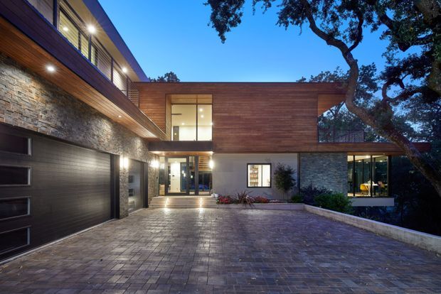 Exterior dusk view of a contemporary multi-level residence arranged around a broad paved driveway or courtyard. The facade combines horizontal timber cladding, rough stacked-stone walls, extensive glazing, recessed lighting, and a large dark garage door. Mature trees frame the property, while warm interior light creates an inviting contrast against the blue evening sky.