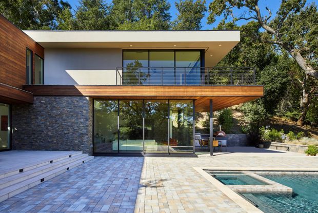 Modern two-storey residence viewed from a paved outdoor terrace and pool area. The house features a flat roof, extensive floor-to-ceiling glazing, warm timber soffits and cladding, a dark stacked-stone wall, and a slender horizontal metal balcony balustrade. Mature trees surround the property, while a swimming pool and broad paved terrace occupy the foreground.