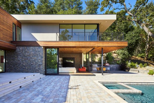 Contemporary two-storey house with a flat roof, extensive glazing, dark stacked-stone walls, and warm timber soffits and cladding. The image looks across a paved pool terrace toward a covered outdoor living area and upper-level balcony, surrounded by mature trees.